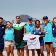 A imagem mostra sete pessoas em pé, ao ar livre, próximas a um corpo d'água, com prédios e uma montanha ao fundo. Elas estão vestidas com roupas esportivas e algumas usam medalhas no pescoço, indicando participação em um evento esportivo. Uma das pessoas segura uma camiseta branca com a palavra "remar" e a imagem de um remo. O ambiente está ensolarado, sugerindo que a foto foi tirada durante o dia.