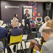 Um homem está em pé falando ao microfone para uma plateia sentada. Uma mulher e um homem também estão no palco, sentados em cadeiras. 