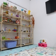 Sala infantil com ambiente acolhedor e brinquedos. À esquerda, há parte de um sofá preto. No centro, uma estante de madeira exibe diversos brinquedos e livros infantis. No chão, um tapete colorido com desenhos de ruas e pistas cobre parte do espaço. À direita, há uma pequena mesa branca com duas cadeiras infantis e brinquedos sobre o tampo. Na parede, acima da estante, há uma televisão preta desligada. O ambiente é bem iluminado, com paredes brancas e aparência organizada e lúdica.