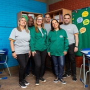 Seis pessoas adultas posam sorridentes dentro de uma sala de aula colorida, com paredes azuis e vermelhas. Três vestem camisetas cinzas e três usam casacos verdes com o logotipo “Educação – Rede Estadual” e a bandeira do Rio Grande do Sul na manga. Ao fundo, há carteiras escolares azuis, cartazes educativos e uma estante com livros.
