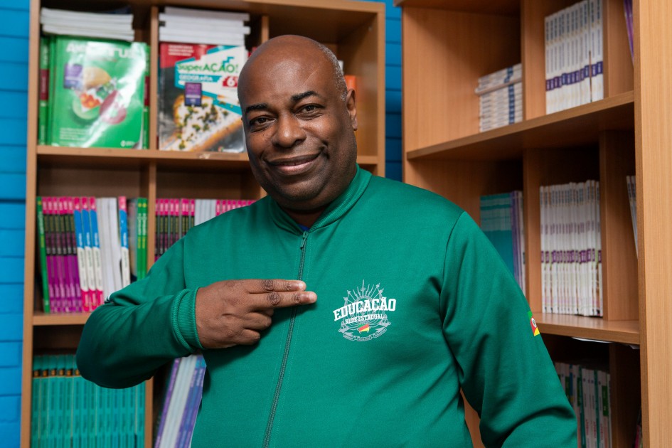 Um homem negro sorri e aponta com a mão para o logotipo “Educação – Rede Estadual” no peito de seu casaco verde. Ele está em frente a uma estante com livros e apostilas organizados. O ambiente parece ser o de uma biblioteca escolar.