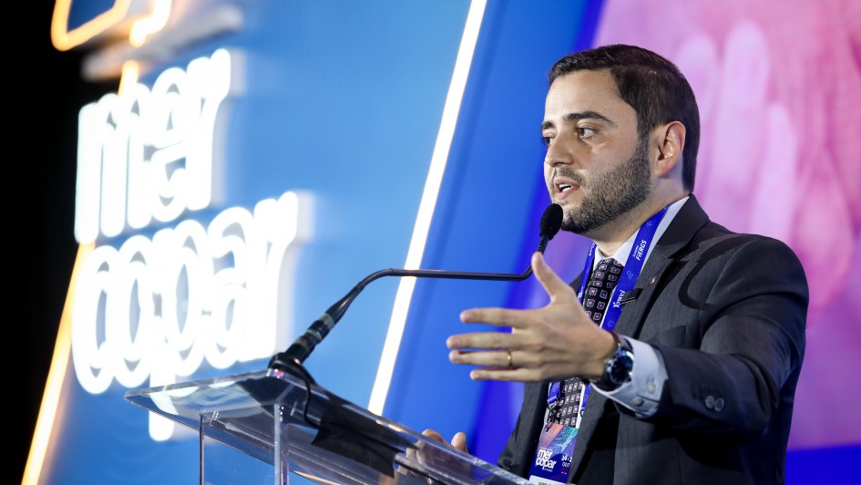 Homem em traje formal fala ao microfone no palco da conferência, com tela iluminada ao fundo exibindo nome do evento. Ambiente profissional e moderno.