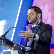 Homem em traje formal fala ao microfone no palco da conferência, com tela iluminada ao fundo exibindo nome do evento. Ambiente profissional e moderno.