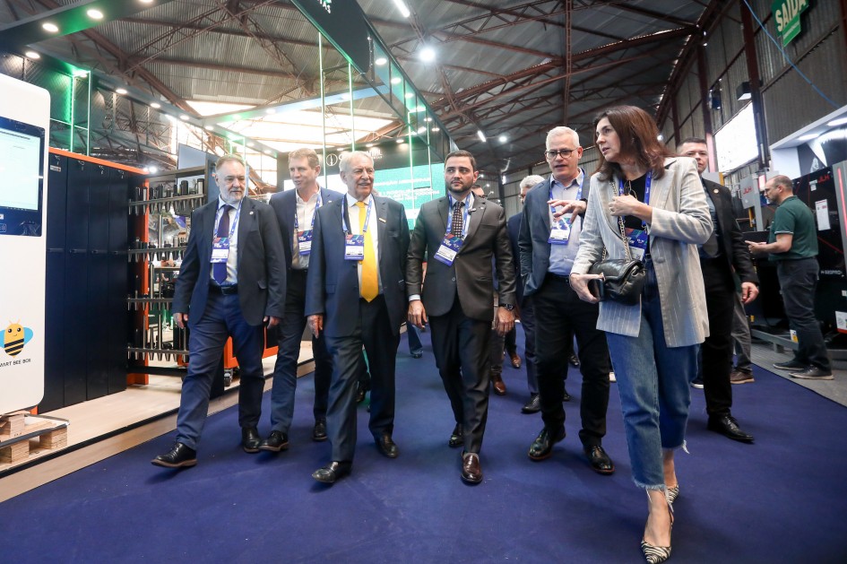 Pessoas caminham juntas em eventos profissionais dentro de um espaço amplo e moderno, com estandes e sinalização. Muitos usam crachás, transferências de feira ou conferência de negócios.