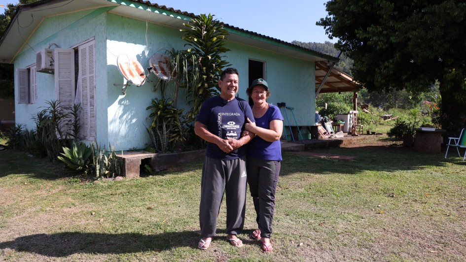 Fotografia de um casal sorridente em frente a uma casa simples de alvenaria pintada de verde-claro, com janelas brancas e telhado de fibrocimento. O homem e a mulher estão de pé sobre a grama, abraçados, ambos vestindo roupas casuais e sandálias. Atrás deles, há plantas próximas à parede da casa e duas antenas parabólicas enferrujadas presas à fachada. Ao fundo, vê-se uma grande árvore, um quintal com utensílios domésticos e vegetação, sob céu azul e ensolarado. A cena transmite um ambiente rural tranquilo.