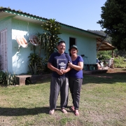 Fotografia de um casal sorridente em frente a uma casa simples de alvenaria pintada de verde-claro, com janelas brancas e telhado de fibrocimento. O homem e a mulher estão de pé sobre a grama, abraçados, ambos vestindo roupas casuais e sandálias. Atrás deles, há plantas próximas à parede da casa e duas antenas parabólicas enferrujadas presas à fachada. Ao fundo, vê-se uma grande árvore, um quintal com utensílios domésticos e vegetação, sob céu azul e ensolarado. A cena transmite um ambiente rural tranquilo.