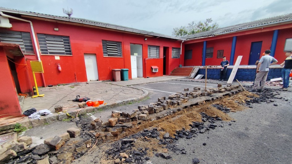 A foto mostra um pátio externo com piso de asfalto parcialmente quebrado, onde há restos de tijolos, areia e pedaços queimados. Ao fundo, há um prédio de paredes vermelhas com portas e janelas retangulares, além de colunas azuis na entrada. Algumas ferramentas e materiais de construção estão espalhados.