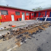 A foto mostra um pátio externo com piso de asfalto parcialmente quebrado, onde há restos de tijolos, areia e pedaços queimados. Ao fundo, há um prédio de paredes vermelhas com portas e janelas retangulares, além de colunas azuis na entrada. Algumas ferramentas e materiais de construção estão espalhados.