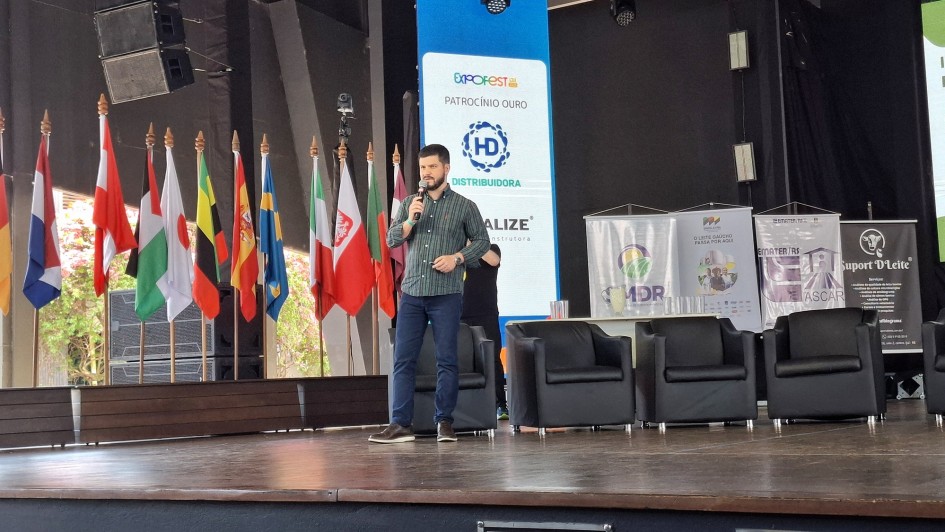 Fotografia de um homem em pé sobre um palco, segurando um microfone e falando ao público durante um evento. Ele veste camisa verde escura e calça jeans. Atrás dele, há diversas bandeiras de países dispostas lado a lado e painéis com logotipos de patrocinadores e instituições, incluindo o da Emater/RS-Ascar e da Secretaria de Desenvolvimento Rural (SDR). À direita, estão dispostas poltronas pretas, indicando espaço para debate ou palestra. O ambiente é iluminado e tem aspecto de auditório ou centro de eventos.