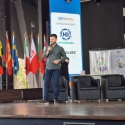 Fotografia de um homem em pé sobre um palco, segurando um microfone e falando ao público durante um evento. Ele veste camisa verde escura e calça jeans. Atrás dele, há diversas bandeiras de países dispostas lado a lado e painéis com logotipos de patrocinadores e instituições, incluindo o da Emater/RS-Ascar e da Secretaria de Desenvolvimento Rural (SDR). À direita, estão dispostas poltronas pretas, indicando espaço para debate ou palestra. O ambiente é iluminado e tem aspecto de auditório ou centro de eventos.