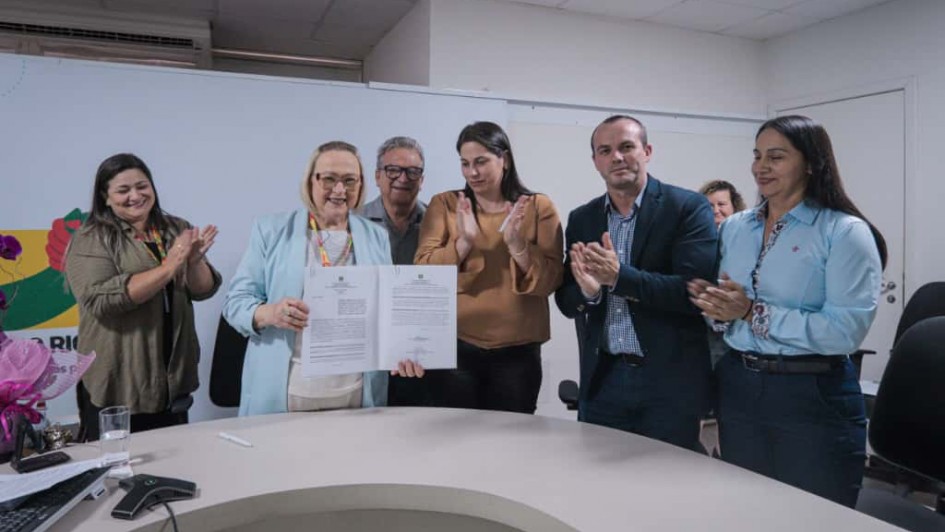 Secretaria Arita Bergmann com documento na mão enquanto outras pessoas na foto batem palmas.