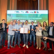 A foto apresenta um grupo de pessoas alinhadas, em frente a um grande painel com a frase “Centro Dia e Avançar Mais SUAS” e logotipos do governo. No centro, duas pessoas seguram um documento aberto. As demais pessoas estão posicionadas lado a lado, algumas com as mãos em gesto de aplauso.