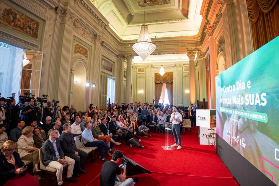 A imagem mostra o governador falando ao microfone, em um púlpito transparente. À esquerda, um público numeroso está sentado em cadeiras, enquanto à direita aparece um grande painel digital com a frase “Centro Dia e Avançar Mais SUAS” e logotipos institucionais.