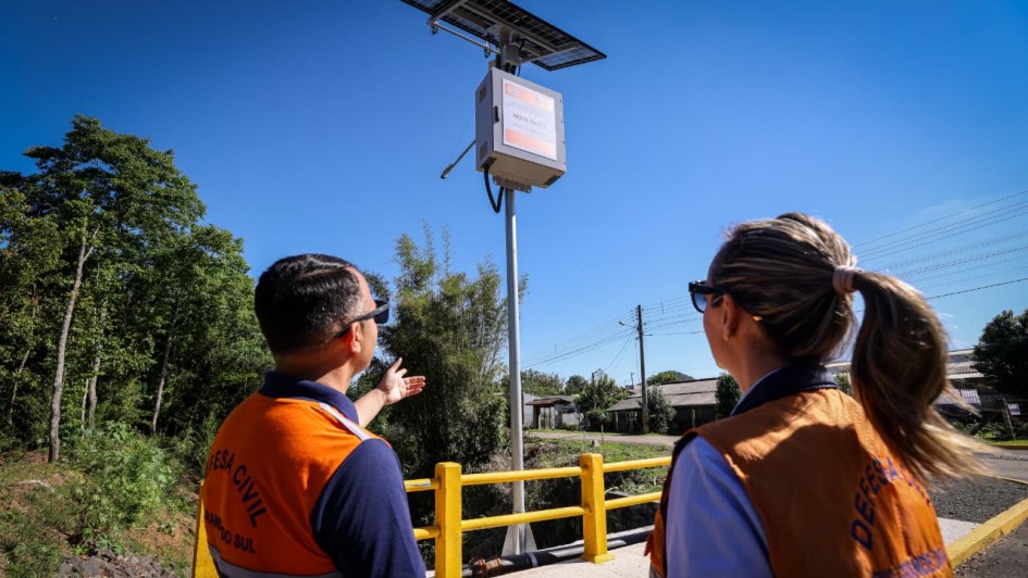 Dois agentes da Defesa Civil, vestindo coletes alaranjados com a inscrição “Defesa Civil – Rio Grande do Sul”, observam e apontam para uma estação de monitoramento recém-instalada em uma ponte. O equipamento possui placa solar e sensores, integrando o sistema de alerta de cheias e eventos climáticos.