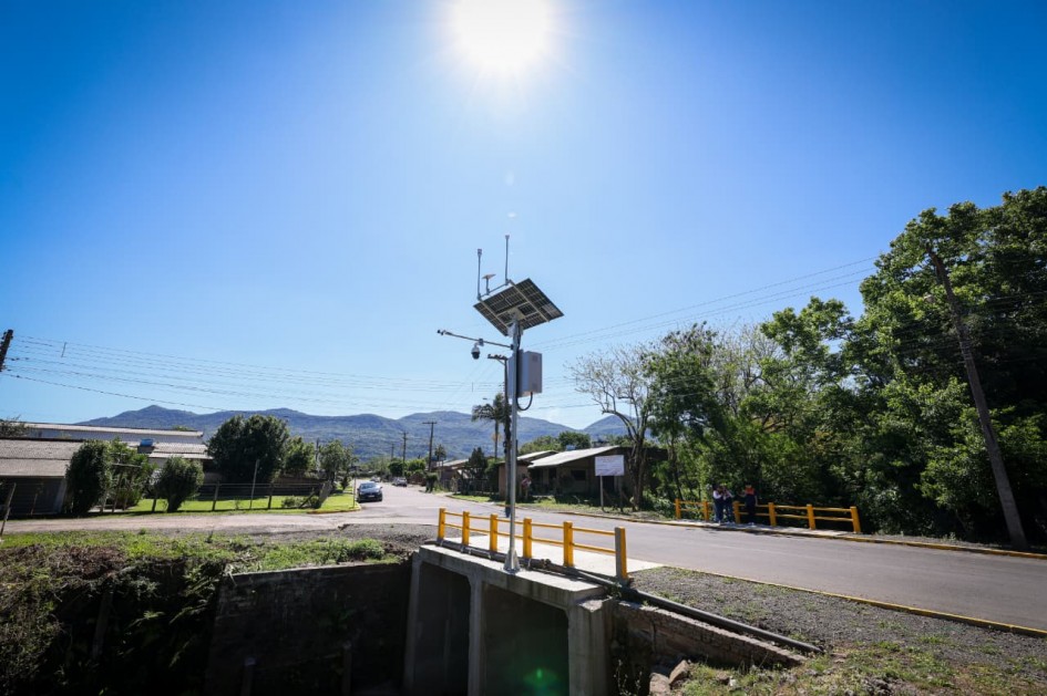 A imagem mostra uma estação hidrometeorológica montada sobre uma ponte, equipada com sensores e painel solar. Ao fundo, aparecem casas e morros sob um céu ensolarado, destacando o ambiente urbano e natural onde o monitoramento será realizado.