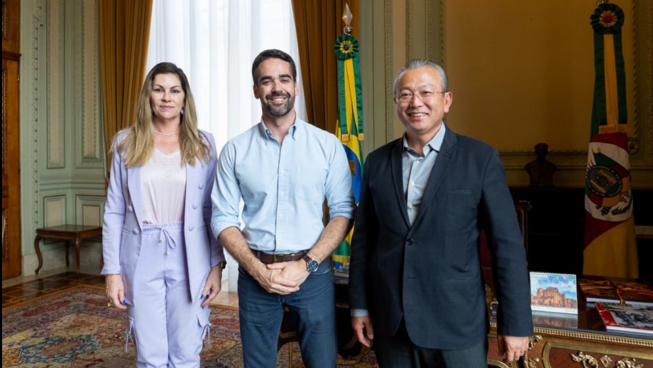 Três pessoas em pé, em um ambiente elegante com cortinas, tapete e móveis de madeira. Atrás delas, há duas bandeiras: a do Brasil e a do Rio Grande do Sul. Parece um escritório de governo ou uma sala oficial.