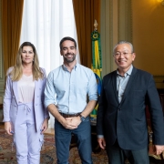 Três pessoas em pé, em um ambiente elegante com cortinas, tapete e móveis de madeira. Atrás delas, há duas bandeiras: a do Brasil e a do Rio Grande do Sul. Parece um escritório de governo ou uma sala oficial.