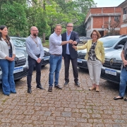 Nove pessoas estão em pé, posando para a foto, ao ar livre, com diversos carros estacionados ao fundo. No centro da imagem, dois homens e uma mulher seguram as chaves de um carro. 