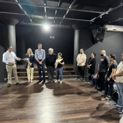 Foto de um palco de teatro, pequeno e rodeado de algumas poucas fileiras de arquibancada. Há 12 pessoas de pé no palco, em roda. 
