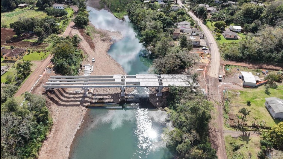 Uma ponte está sendo construída sobre um rio em uma área rural. Há estruturas metálicas, máquinas e materiais nos dois lados do rio. Casas e vegetação cercam o local, e a obra conecta duas partes da comunidade.