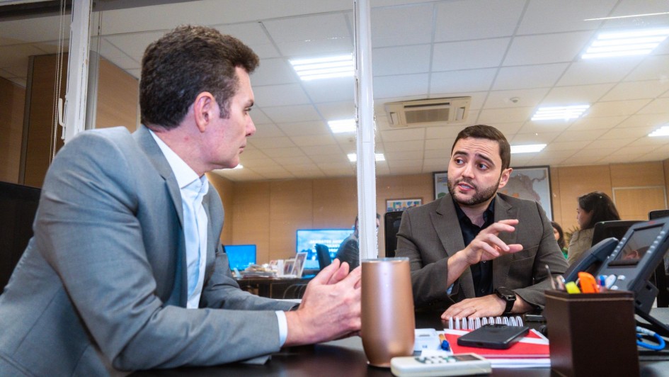 Dois homens sentados em uma mesa de escrit&oacute;rio conversam, em ambiente iluminado por luzes de teto. O homem &agrave; esquerda veste terno cinza e camisa clara, enquanto o da direita usa terno escuro e gesticula com a m&atilde;o durante a fala. Sobre a mesa, h&aacute; objetos de escrit&oacute;rio, como canetas, caderno, telefone e uma garrafa t&eacute;rmica. Ao fundo, outras pessoas trabalham em suas mesas.