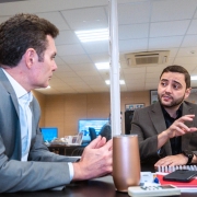 Dois homens sentados em uma mesa de escritório conversam, em ambiente iluminado por luzes de teto. O homem à esquerda veste terno cinza e camisa clara, enquanto o da direita usa terno escuro e gesticula com a mão durante a fala. Sobre a mesa, há objetos de escritório, como canetas, caderno, telefone e uma garrafa térmica. Ao fundo, outras pessoas trabalham em suas mesas.