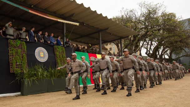 Governador Eduardo Leite participa da formatura de novos soldados da Brigada Militar. Na foto, ele está no palanque oficial com outras autoridades. Em primeiro plano, uma tropa de policiais militares passa marchando.