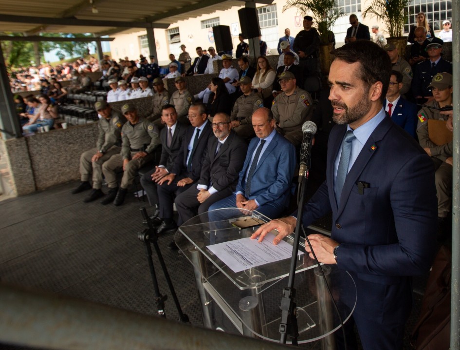 O governador Eduardo Leite discursa ao microfone em um púlpito de acrílico, durante a cerimônia de formatura. Atrás dele, autoridades civis e militares acompanham a solenidade.