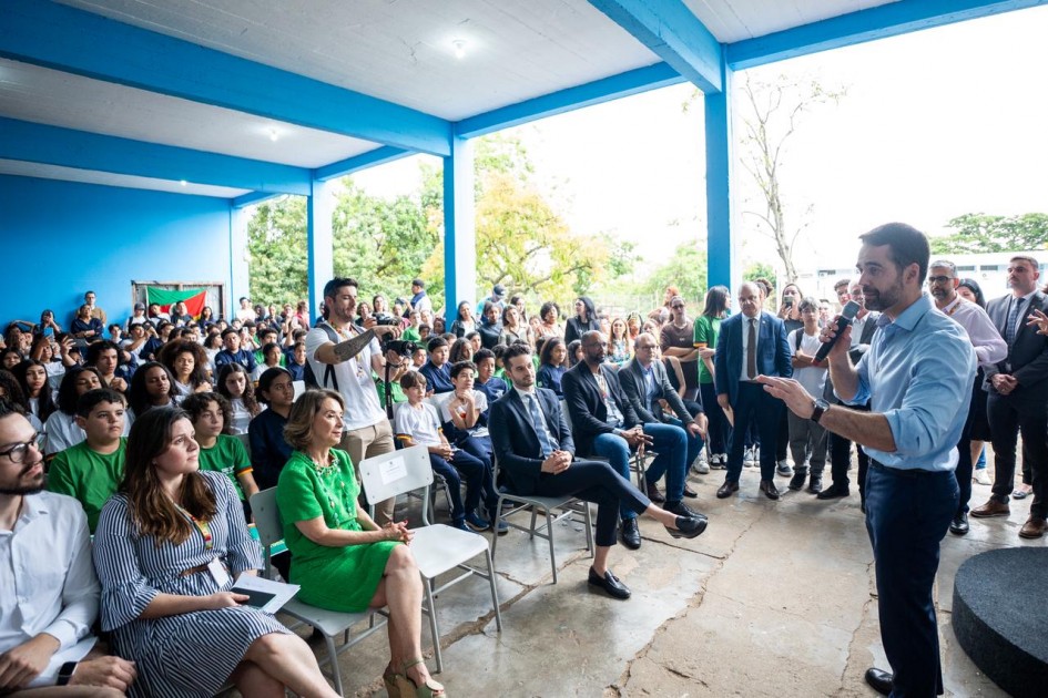 A foto mostra um espaço coberto, onde ocorre um evento escolar com grande público. No primeiro plano, o governador está falando ao microfone, voltado para um grupo sentado em cadeiras, incluindo autoridades e convidados. Ao fundo, há dezenas de estudantes uniformizados.
