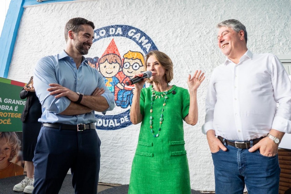 A foto mostra dois homens e uma mulher em pé conversando diante de uma parede branca com um grande logotipo circular que contém ilustrações de crianças e o texto “Paulo da Gama”. A mulher fala ao microfone e gesticula.