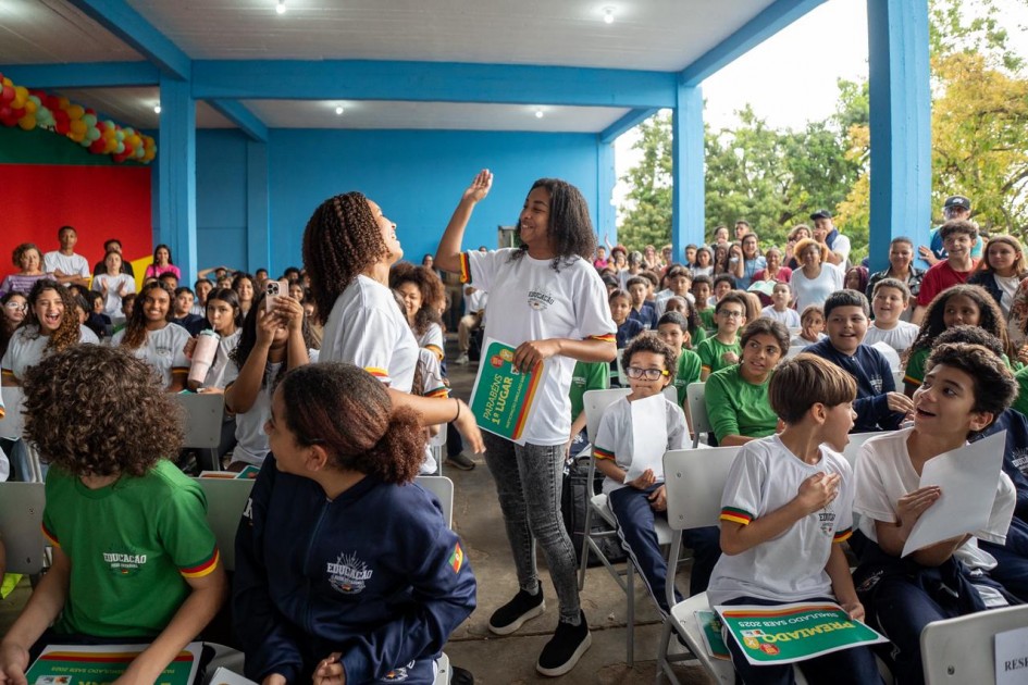 A imagem apresenta um grupo de estudantes sentados em cadeiras organizadas em fileiras. Duas estudantes estão em pé, interagindo entre si, segurando cartazes verdes com a inscrição “Premiado Simulado Saeb 2025”.