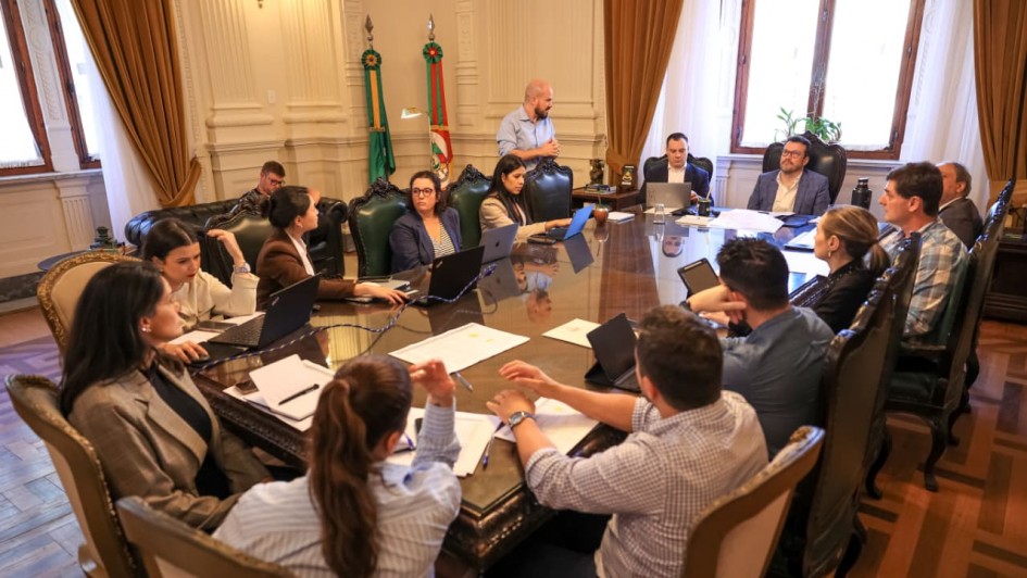 Em uma sala ampla e bem iluminada, com cortinas douradas e bandeiras do Rio Grande do Sul ao fundo, um grupo de pessoas está reunido ao redor de uma grande mesa de madeira. Elas utilizam notebooks, papéis e canetas, demonstrando concentração em uma reunião de trabalho. Um homem em pé fala ao grupo, enquanto os demais escutam atentamente. O ambiente possui decoração clássica e mobiliário elegante.