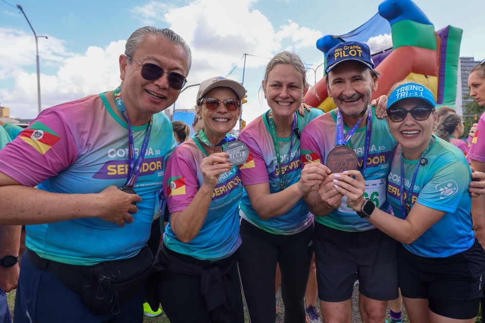 Cinco pessoas sorridentes exibem medalhas após corrida, vestem camisetas coloridas com nome do evento. Ao fundo, arco inflável e clima de celebração.