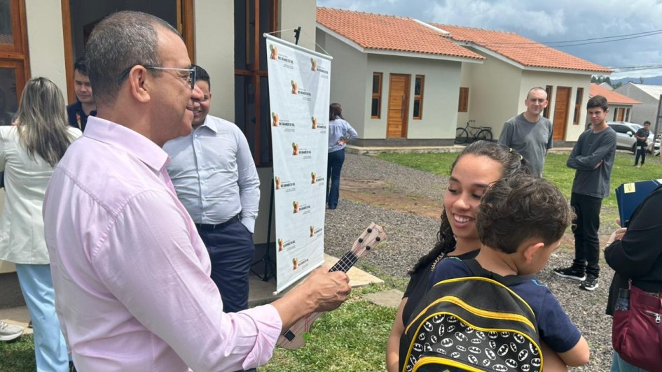 Homem entrega chave a mulher com criança no colo, em frente a casas novas. Cena transmite emoção e conquista, possivelmente entrega de moradia em evento comunitário.