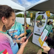 Duas pessoas interagem em estande de evento ao ar livre. Uma segurança celular, outra exibe cartaz com QR code e frase “Servidor Público”. Ambiente com tendas e clima de campanha.