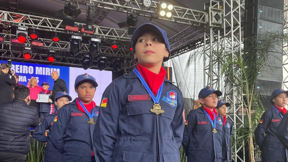 A imagem mostra crianças em forma, perfiladas e uniformizadas em tom azul marinho com um lenço vermelho. Elas estão com medalhas no peito e posicionadas sob uma estrutura de metal com um palco ao fundo.