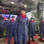 A imagem mostra crianças em forma, perfiladas e uniformizadas em tom azul marinho com um lenço vermelho. Elas estão com medalhas no peito e posicionadas sob uma estrutura de metal com um palco ao fundo. 