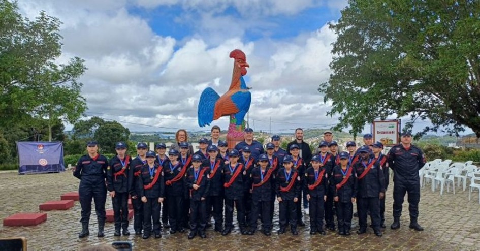 Imagem ao ar livre mostra um grupo de crianças e alguns adultos em pé, posicionados lado a lado para a foto. As crianças e os adultos vestem uniformes de bombeiros militares, em tom azul marinho.