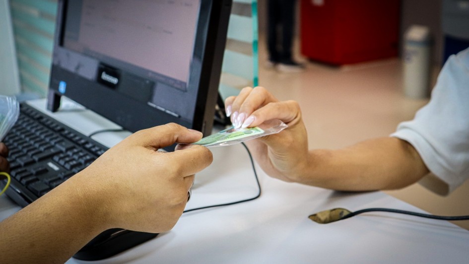 Foto de uma banca de atendimento, com um computador ao fundo. Em destaque está uma mão entregando uma carteira de identidade a outra, que pega o documento.