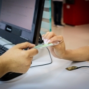 Foto de uma banca de atendimento, com um computador ao fundo. Em destaque está uma mão entregando uma carteira de identidade a outra, que pega o documento.