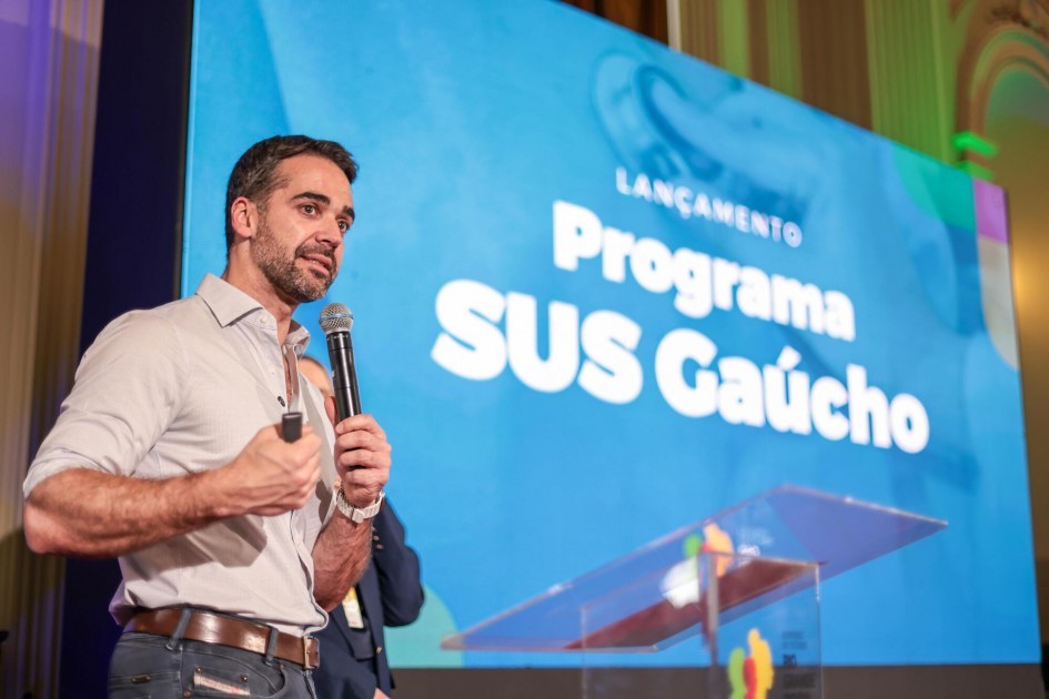 Foto do governador, de pé, segurando um microfone e falando em frente a um telão azul que mostra as palavras “Programa SUS Gaúcho”.