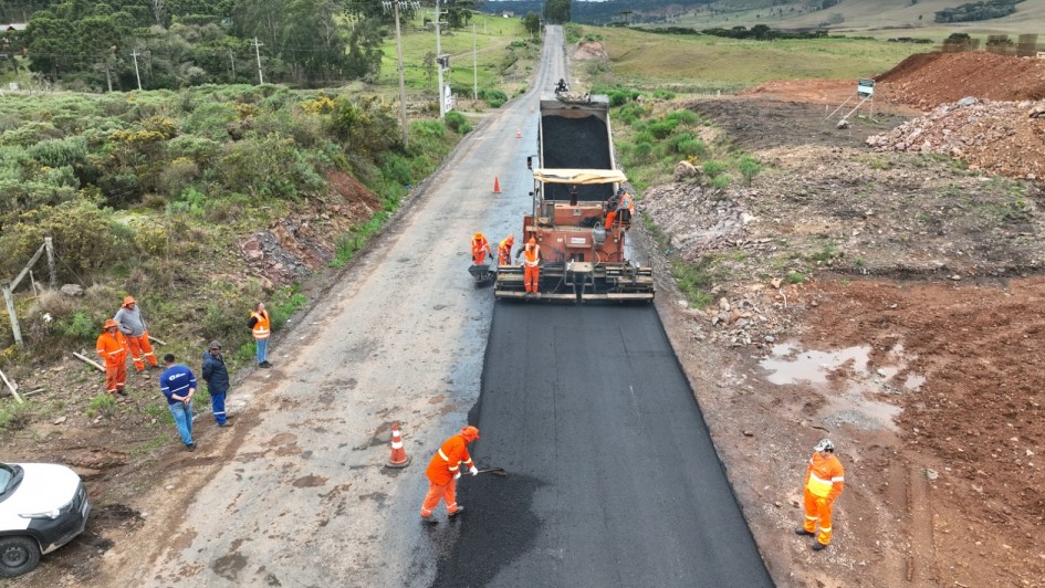 Imagem mostra um grupo de trabalhadores com uniformes laranja realizando obras de pavimentação asfáltica em uma rodovia. Uma máquina espalha a camada de asfalto novo enquanto outros operários trabalham manualmente na aplicação e nivelamento do material. À esquerda, alguns trabalhadores observam a execução do serviço, próximos a um veículo branco. A estrada está cercada por vegetação e áreas de terra, em uma região de relevo levemente ondulado.