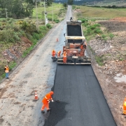 Imagem mostra um grupo de trabalhadores com uniformes laranja realizando obras de pavimentação asfáltica em uma rodovia. Uma máquina espalha a camada de asfalto novo enquanto outros operários trabalham manualmente na aplicação e nivelamento do material. À esquerda, alguns trabalhadores observam a execução do serviço, próximos a um veículo branco. A estrada está cercada por vegetação e áreas de terra, em uma região de relevo levemente ondulado.