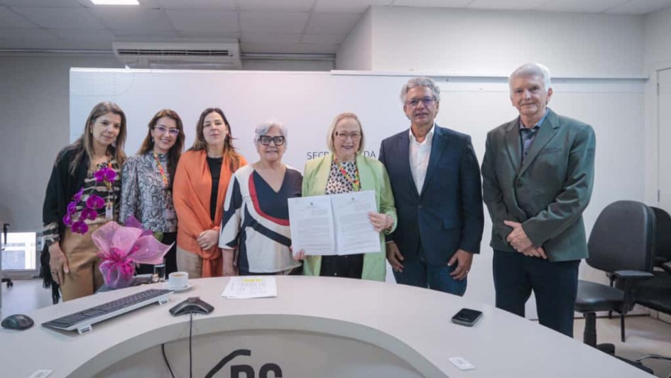 Foto posada em ambiente fechado de cor branca. Cinco mulheres e dois homens aparecem de pé, de frente para a câmera, em trajes formais, sorrindo. No meio, Arita segura e exibe o documento assinado.