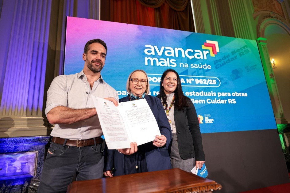 Foto posada de três pessoas, de pé, em ambiente fechado. Leite, ao lado de Arita e outra mulher, segura e exibe um documento assinado. Atrás deles, há um telão colorido com os dizeres “Avançar mais na saúde”.