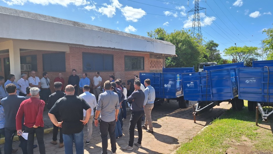 Em área externa, um grupo de cerca de 20 pessoas acompanha uma cerimônia em frente a um prédio de tijolos, sob céu azul e ensolarado. À direita da imagem, há carretas agrícolas azuis enfileiradas, que fazem parte dos equipamentos entregues pelo governo do Estado. Algumas autoridades discursam enquanto os participantes observam.