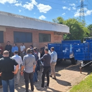 Em área externa, um grupo de cerca de 20 pessoas acompanha uma cerimônia em frente a um prédio de tijolos, sob céu azul e ensolarado. À direita da imagem, há carretas agrícolas azuis enfileiradas, que fazem parte dos equipamentos entregues pelo governo do Estado. Algumas autoridades discursam enquanto os participantes observam.