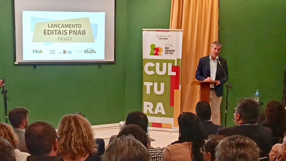 Homem em pé fala ao microfone em um púlpito de madeira, diante de uma plateia sentada em auditório. Ao fundo, há um telão com o texto “Lançamento Editais PNAB Ciclo 2” e, ao lado, um banner da Secretaria da Cultura do Rio Grande do Sul com a palavra “CULTURA” em destaque. O ambiente tem paredes verdes e cortina amarela.