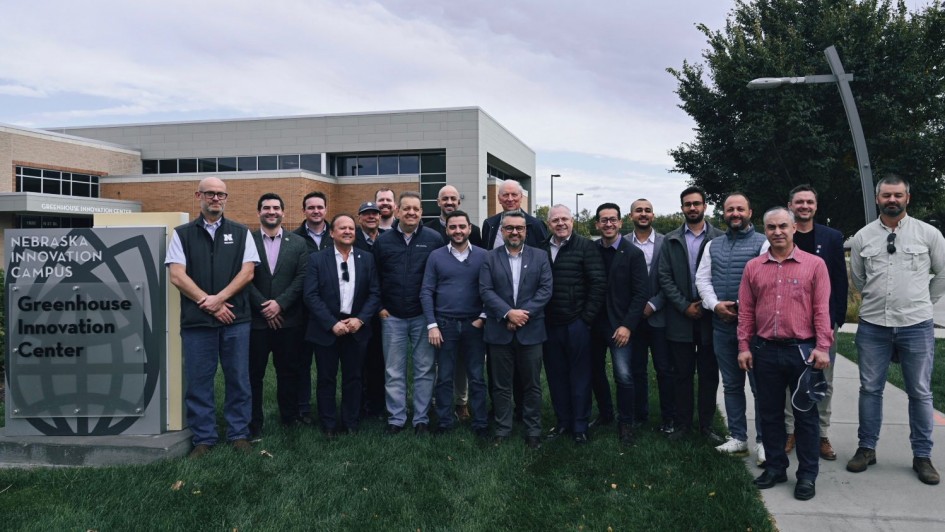 Uma foto externa de um grupo de cerca de 20 homens de pé, posando na frente de um letreiro e um prédio. O letreiro de cor escura, no canto inferior esquerdo, diz "NEBRASKA INNOVATION CAMPUS" e, abaixo, "Greenhouse Innovation Center". O prédio atrás é moderno, de dois andares, com fachada em cores claras e muitas janelas. Os homens estão vestidos de maneira casual de negócios ou casual e parecem ser membros de uma comitiva, possivelmente na área de agricultura ou tecnologia de estufas, em visita ao centro de pesquisa no Nebraska, EUA. O céu está nublado.
