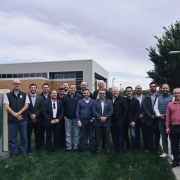 Uma foto externa de um grupo de cerca de 20 homens de pé, posando na frente de um letreiro e um prédio. O letreiro de cor escura, no canto inferior esquerdo, diz "NEBRASKA INNOVATION CAMPUS" e, abaixo, "Greenhouse Innovation Center". O prédio atrás é moderno, de dois andares, com fachada em cores claras e muitas janelas. Os homens estão vestidos de maneira casual de negócios ou casual e parecem ser membros de uma comitiva, possivelmente na área de agricultura ou tecnologia de estufas, em visita ao centro de pesquisa no Nebraska, EUA. O céu está nublado.